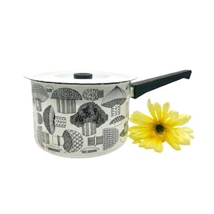 May include: White enamel saucepan with a black mushroom pattern and a black handle. The pot has a white lid with a black knob. A yellow flower is next to the pot. Kitchenware.