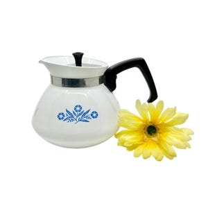 May include: White vintage coffee pot with a black handle and lid. The pot features a blue floral design and a silver band around the middle. A yellow flower with a green center sits next to the pot.