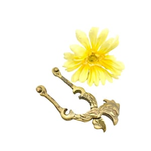May include: A gold-toned brass nutcracker with an ornate design. The nutcracker is shaped like a bird with its wings spread out. The nutcracker is sitting on a white background next to a yellow flower.