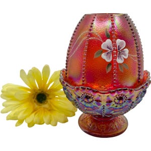 May include: An iridescent red glass egg-shaped decorative piece with a floral design and beaded accents. The piece sits in a matching pedestal base. A yellow flower is next to the decorative piece.