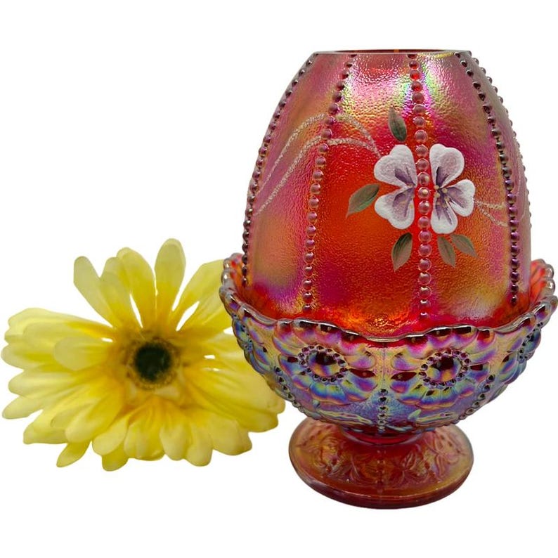May include: An iridescent red glass egg-shaped decorative piece with a floral design and beaded accents. The piece sits in a matching pedestal base. A yellow flower is next to the decorative piece.