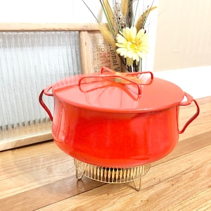 May include: A vintage red enamel pot with a matching lid and handles. The pot sits on a gold-colored metal stand. The background includes a wooden framed window and a vase of flowers.