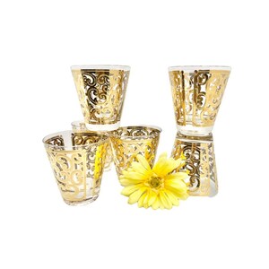 May include: Set of five clear glass tumblers with gold-colored intricate designs. The glasses are arranged in a pyramid shape with a yellow flower in front.