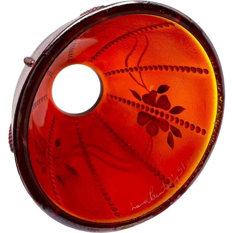 May include: A hand-painted, red glass lampshade with a floral design. The lampshade has a cylindrical shape with a large circular opening. The floral design is dark red and includes a flower and leaves. The words "hand painted by Stan" are written on the lampshade.
