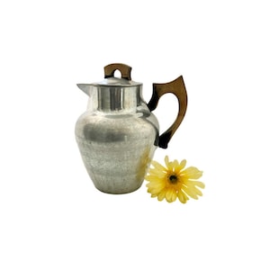 May include: A silver pitcher with a hammered finish and brown wooden handles. A yellow flower is in the foreground.