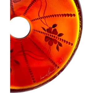 May include: Close-up of a decorative, orange glass plate with a central hole. The plate features hand-painted floral and dot patterns in red and brown. The edge is outlined in yellow.
