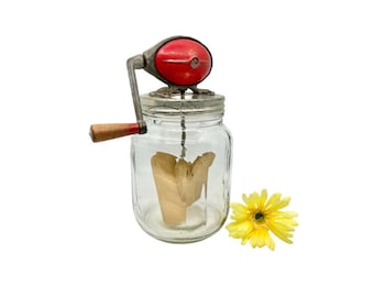 May include: Vintage butter churn with a glass jar and a red and silver crank. The churn is filled with a wooden paddle and a yellow flower is in the foreground.