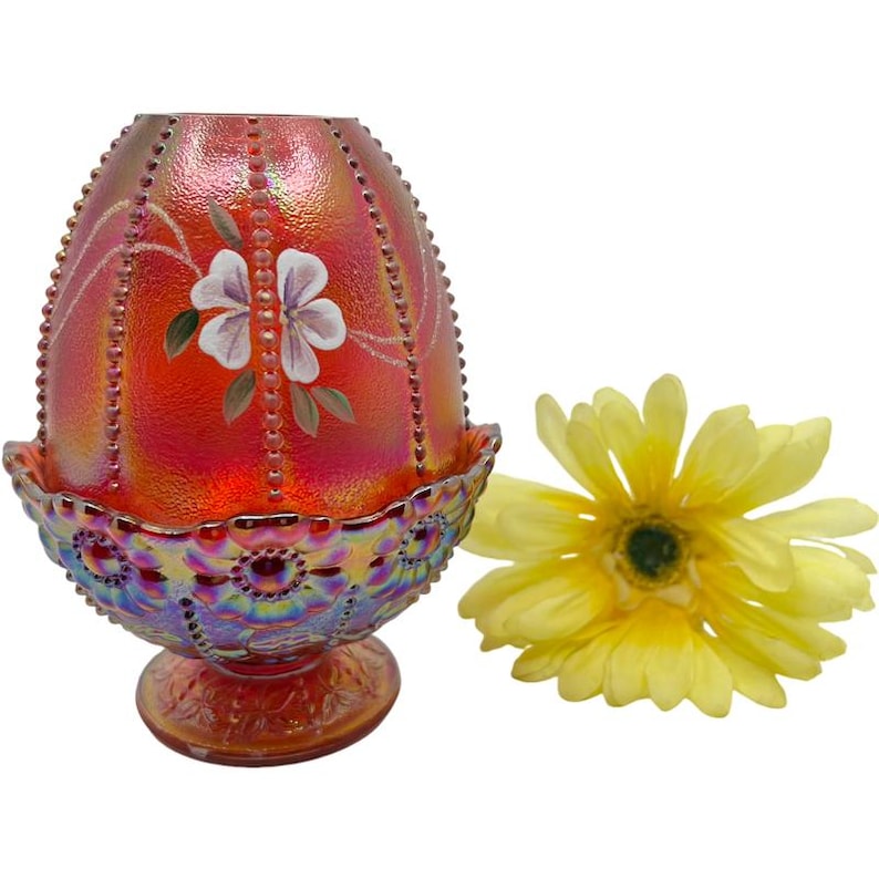 May include: An iridescent red and orange egg-shaped decorative glass piece with a floral design and a yellow flower. The glass piece has a beaded border and sits on a decorative base.