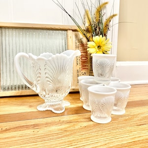 May include: A set of six white milk glass cups and a matching pitcher with a scalloped edge and a seashell design.