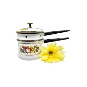 May include: White enamel double boiler with black handles and a lid. The pot features a fruit and floral design with gold trim. A yellow gerbera daisy is next to the pot.