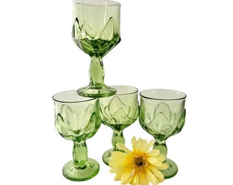 May include: Set of four green glass wine glasses with a textured design. The glasses have a tall, slender stem and a wide bowl. A yellow flower is in front of the glasses.