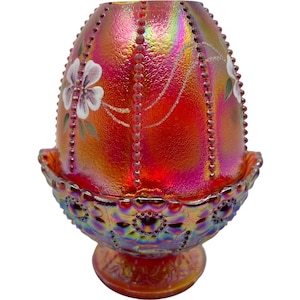 May include: An iridescent red glass egg-shaped decorative piece with a matching base. The egg has a textured surface with vertical beaded accents and painted white flowers. The base is also iridescent with a detailed pattern.
