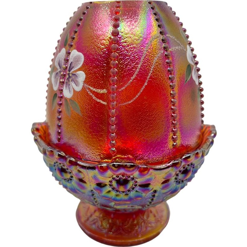May include: An iridescent red glass egg-shaped decorative piece with a matching base. The egg has a textured surface with vertical beaded accents and painted white flowers. The base is also iridescent with a detailed pattern.