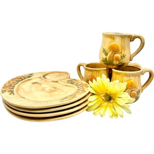 May include: Set of four ceramic mushroom-themed mugs and four matching plates. The mugs and plates are decorated with brown, yellow, and green mushrooms.