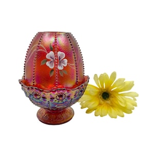 May include: A red iridescent glass candle holder with a floral design. The candle holder has a decorative base and is adorned with painted white flowers. A yellow gerbera daisy is next to the candle holder.