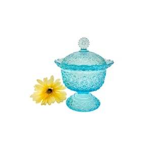 May include: A blue glass candy dish with a scalloped edge and a floral design. The dish has a lid with a flower knob. A yellow flower is in the foreground.