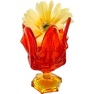 May include: A red and orange glass vase with a ruffled top and a hexagonal base. A yellow flower is in the vase.