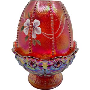 May include: A vibrant red iridescent glass egg-shaped decorative piece. The egg features a hand-painted white and purple flower design with green leaves. The egg sits in a matching iridescent dish with a decorative base.