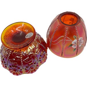May include: Two iridescent orange glass bowls. One bowl is upside down, revealing a detailed pattern on the base. The other bowl is upright, with a floral design and white painted flowers. The bowls have a textured surface and are likely decorative.