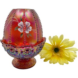 May include: A red iridescent glass egg-shaped decorative container with a floral design. The container has a beaded rim and sits on a decorative base. A yellow flower is next to the container.