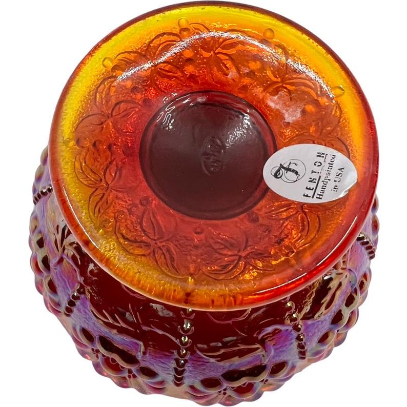 May include: An overhead view of a hand-painted Fenton glass bowl. The bowl is iridescent with a red and orange color scheme. The rim has a floral pattern, and the base is a deep red. A sticker on the side reads "Fenton Handpainted in USA."
