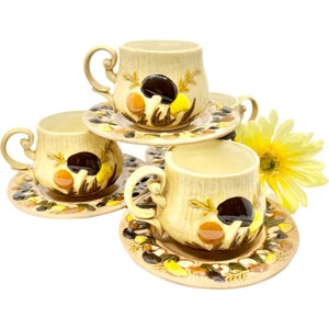 May include: Set of four vintage ceramic teacups and saucers with a mushroom design. The cups and saucers are beige with brown, orange, and yellow mushrooms.