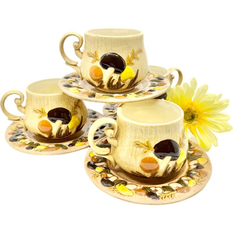 May include: Set of four vintage ceramic teacups and saucers with a mushroom design. The cups and saucers are beige with brown, orange, and yellow mushrooms.