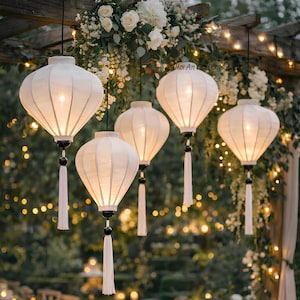 May include: Five white paper lanterns with black accents and white tassels hang from a wooden structure decorated with white flowers and string lights. The lanterns are lit, creating a warm glow.