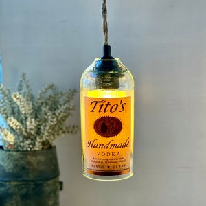 May include: A clear glass bottle with a light inside, hanging from a rope. The bottle is labeled with "Tito's Handmade Vodka" and the text "Award Winning American Vodka Crafted in Old Fashioned Pot Still Austin, Texas".