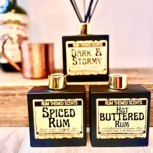 May include: Two black square glass bottles with gold lids and labels. The labels are gold with black text. The left bottle says "RUM THEMED SCENTS SPICED RUM" and the right bottle says "RUM THEMED SCENTS HOT BUTTERED RUM".
