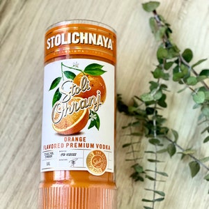 以下が含まれることがあります： ストリ・オランジ オレンジフレーバープレミアムウォッカのガラス瓶。ボトルはオレンジ色で、ストリロゴと「ストリ・オランジ」の文字が金色のフォントで書かれたラベルが付いています。ボトルは木製の表面の上に置かれています。