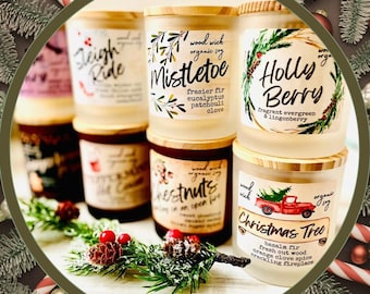May include: A collection of Christmas-themed candles with wooden lids. The labels feature festive text like "Mistletoe," "Holly Berry," and "Christmas Tree," along with illustrations of a red truck and evergreen branches. The candles are arranged with holiday decorations.