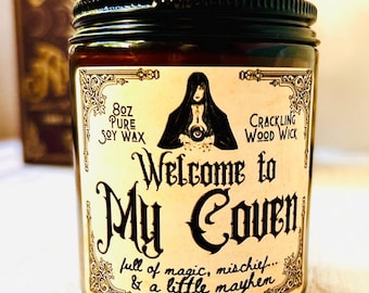 May include: A dark amber glass jar with a black lid, featuring a cream-colored label. The label reads "Welcome to My Coven" with a witch illustration. The jar contains 8 oz of pure soy wax with a crackling wood wick.