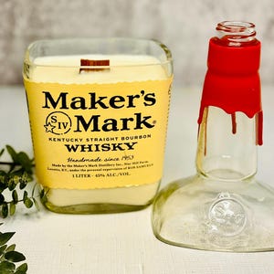 May include: A Maker's Mark whisky candle in a repurposed bottle, with a wooden wick. The candle has a yellow label with black text. The empty bottle has a red wax seal. The candle is handmade since 1953.