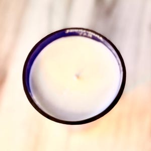 May include: A white candle with a blue rim in a glass jar. The candle is unlit and has a small indentation in the center.