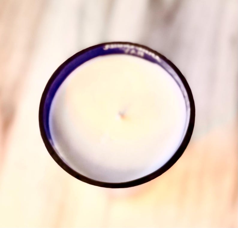 May include: A white candle with a blue rim in a glass jar. The candle is unlit and has a small indentation in the center.