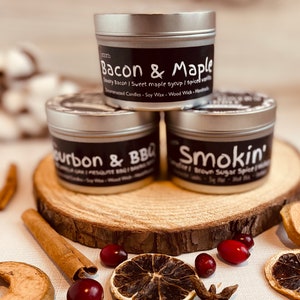 以下が含まれることがあります： 黒いラベルが付いた銀色のキャンドル缶が3つ。ラベルには、「Bacon & Maple」、「Bourbon & BBQ」、「Smokin' Brown Sugar Spice」という白い文字が書かれています。キャンドルは丸い木の板の上に並べてあります。