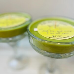 May include: Two green glass margarita glasses filled with green scented candles. The candles have a label that reads "Deconstructed Candles" and "www.deconstructedcandles.com".