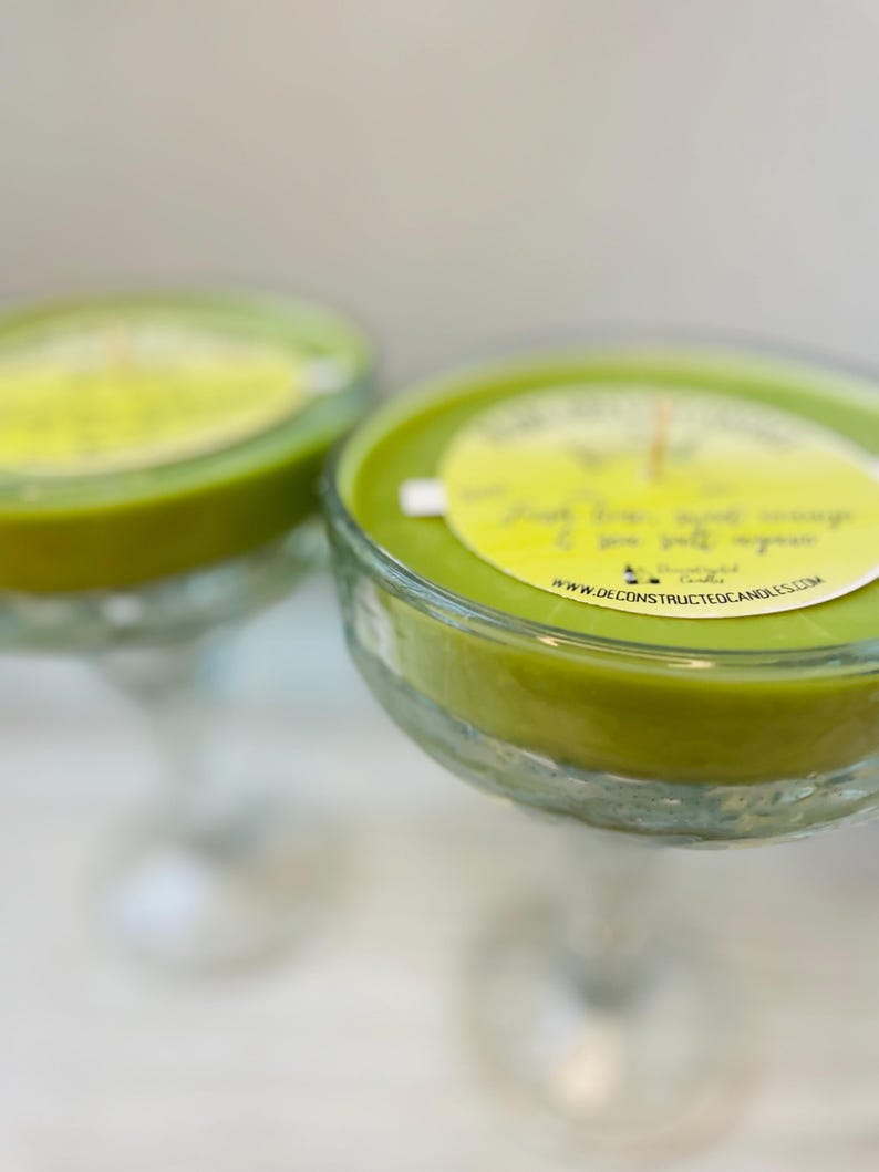 May include: Two green glass margarita glasses filled with green scented candles. The candles have a label that reads "Deconstructed Candles" and "www.deconstructedcandles.com".