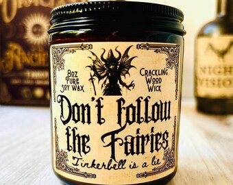 May include: A dark glass jar with a black lid and a label that reads "Don't follow the Fairies, Tinkerbell is a lie." The label features a winged creature illustration and text indicating 8 oz of pure soy wax and a crackling wood wick.