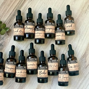 May include: A collection of 14 small brown glass bottles with droppers, each labeled with a different scent name. The bottles are arranged in two rows on a light brown wooden surface. The scents include French Rose, Pomelo, Vintage Island, Champagne, Lemon Drop, French 75, Bourbon, Margarita, On the Beach, Lavender Garden, Boho, Old Fashioned, Sea Witch, and Red Sangria.