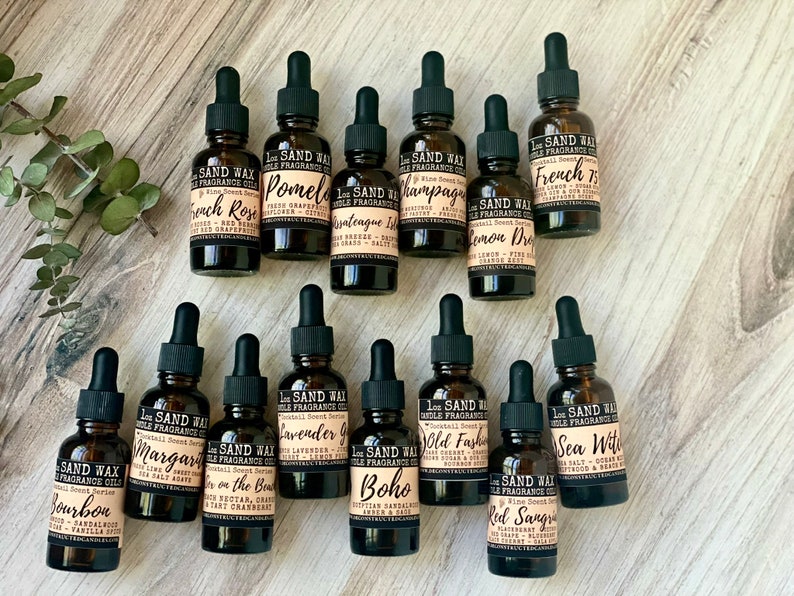 May include: A collection of 14 small brown glass bottles with droppers, each labeled with a different scent name. The bottles are arranged in two rows on a light brown wooden surface. The scents include French Rose, Pomelo, Vintage Island, Champagne, Lemon Drop, French 75, Bourbon, Margarita, On the Beach, Lavender Garden, Boho, Old Fashioned, Sea Witch, and Red Sangria.