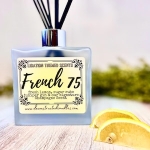 May include: A square, frosted glass diffuser bottle with black reeds. The label reads "Libation Themed Scents French 75" with scent notes. Two lemon slices are in the foreground. The website www.deconstructedcandles.com is also visible.