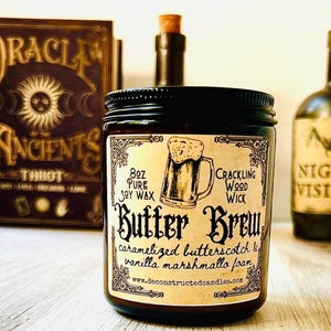K&ouml;nnte beinhalten: Eine dunkle Glas-Kerze im Glas mit schwarzem Deckel, beschriftet mit "Butter Brew" mit einer Bierkrug-Illustration. Das Etikett lautet auch "8oz Pure Soy Wax, Crackling Wood Wick, karamellisierter Butterscotch & Vanille-Marshmallow-Schaum". Ein Tarotkartenbuch und zwei Flaschen sind im Hintergrund.