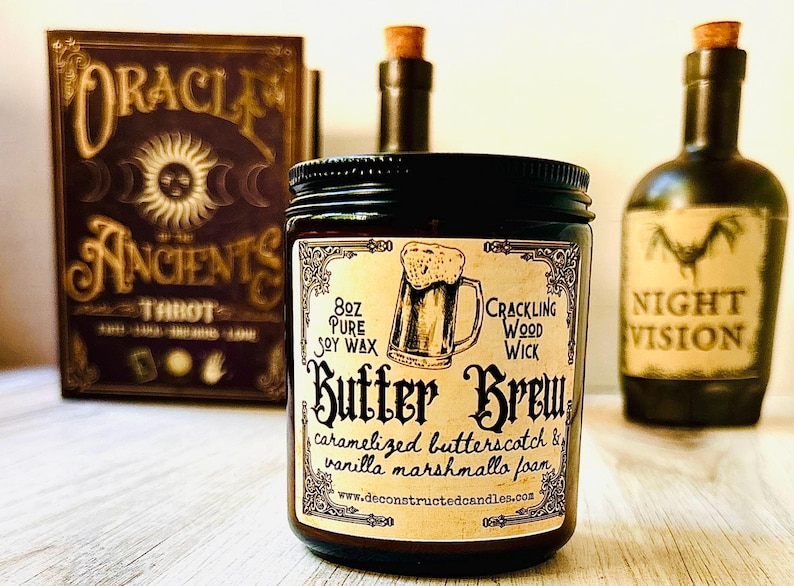 K&ouml;nnte beinhalten: Eine dunkle Glas-Kerze im Glas mit schwarzem Deckel, beschriftet mit "Butter Brew" mit einer Bierkrug-Illustration. Das Etikett lautet auch "8oz Pure Soy Wax, Crackling Wood Wick, karamellisierter Butterscotch & Vanille-Marshmallow-Schaum". Ein Tarotkartenbuch und zwei Flaschen sind im Hintergrund.