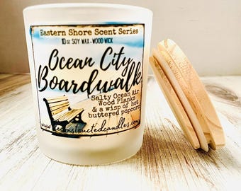 May include: A frosted glass candle with the text "Ocean City Boardwalk" and a wooden wick. The label includes a bench illustration and scent notes. A wooden candle lid is next to the candle.