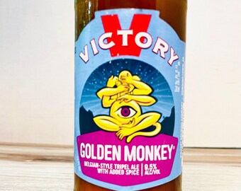 May include: A brown glass beer bottle with a blue and red label. The label features the word "VICTORY" and a yellow monkey-like figure with a single eye. The words "GOLDEN MONKEY" are also on the label, along with text indicating it is a Belgian-style tripel ale.