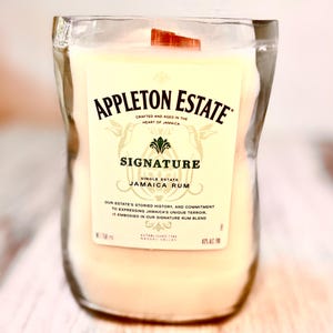 Pode incluir: Uma vela branca em uma garrafa de vidro transparente com uma tampa de madeira marrom. A garrafa tem um rótulo que diz "Appleton Estate Signature Single Estate Jamaica Rum". O rótulo também inclui o texto "Our estate's storied history, and commitment to expressing Jamaica's unique terroir, is embodied in our signature rum blend." e "Established 1749 Nassau Valley".