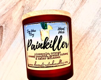 May include: A 10 oz soy wax candle with a wood wick in a clear glass jar with a wooden lid. The candle is labeled "Painkiller" and has a tropical scent with notes of pineapple, orange, creamy coconut, grated nutmeg, and amber molasses. The label also includes the website "deconstructedcandles.com".