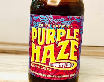 May include: A brown glass bottle of Abita Brewing Purple Haze Raspberry Lager. The label features the words "Purple Haze" in large yellow letters, with a purple and red background. The bottle contains 4.2% alcohol by volume.